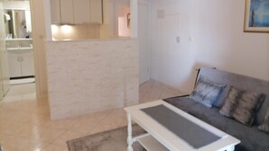 Una sala de estar con un sofá gris, una mesa de centro blanca y una zona de cocina con gabinetes blancos y un salpicadero de azulejos.