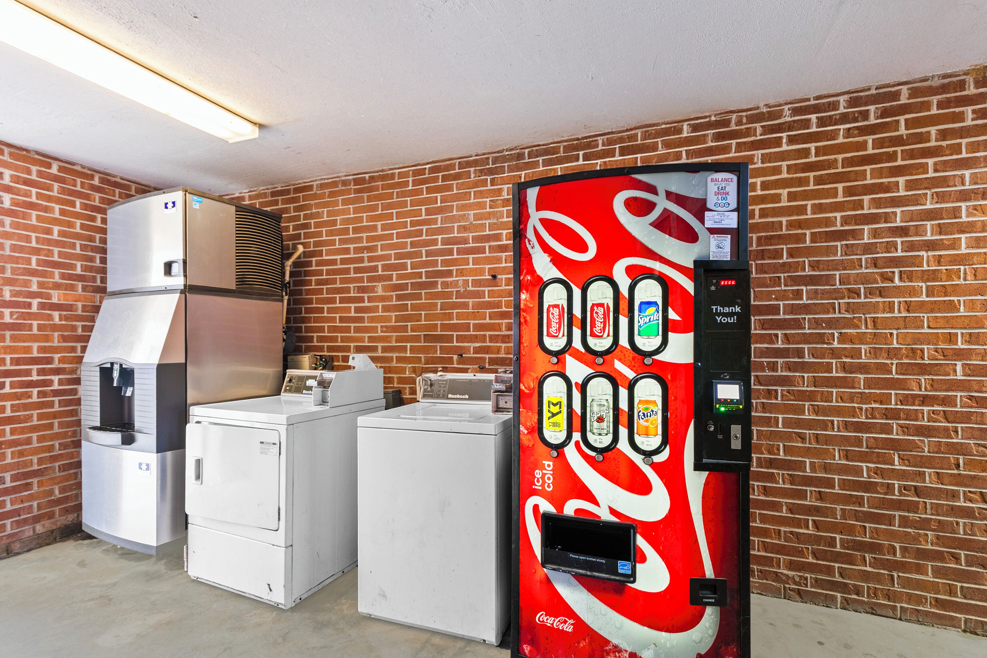 A vending machine with a Coca-Cola logo, a water dispenser, and a coffee machine in a room with brick walls and a concrete floor.