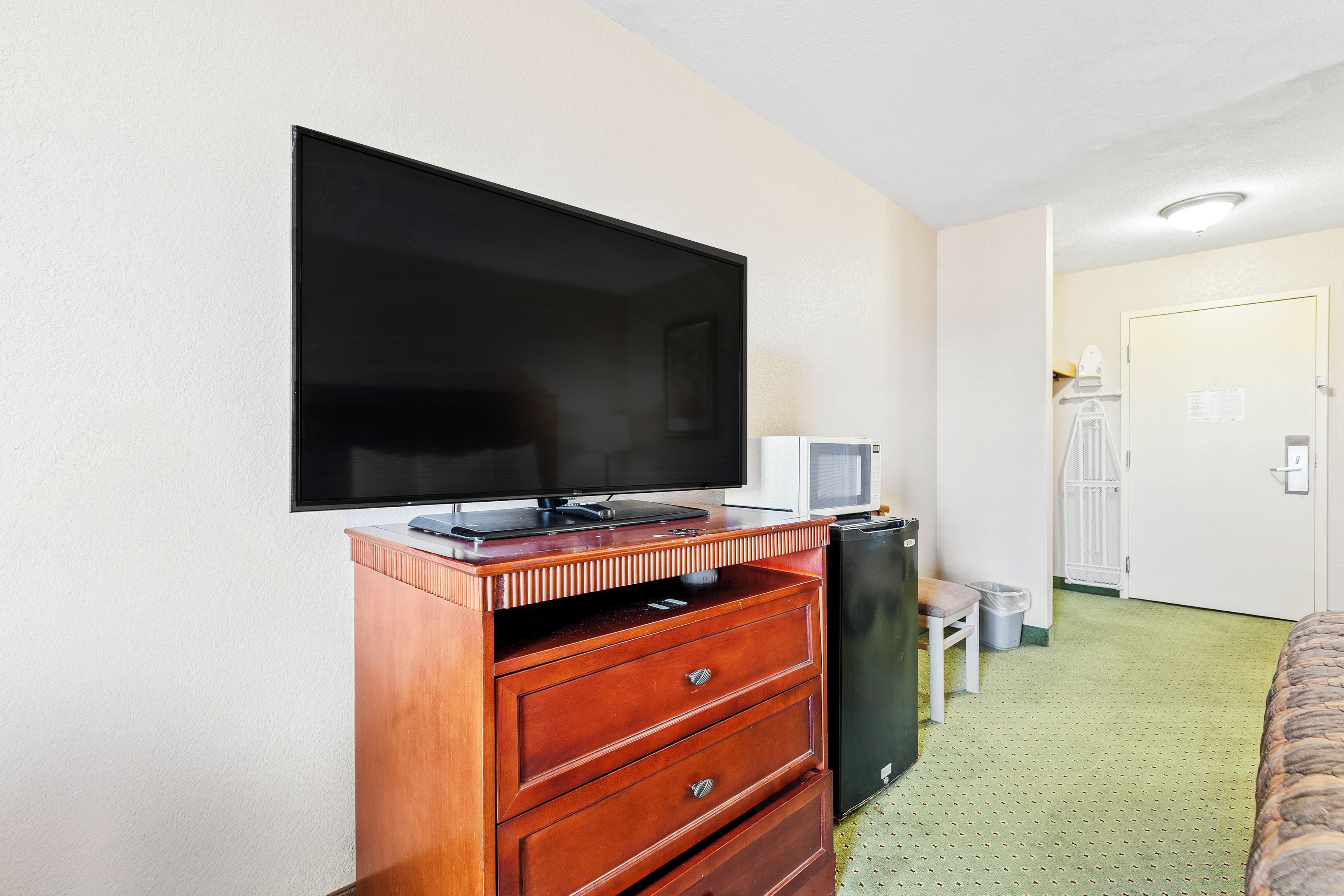 Une chambre d’hôtel équipée d’un téléviseur à écran plat fixé à un meuble en bois, d’une petite table avec une lampe et d’une porte donnant sur une autre pièce.