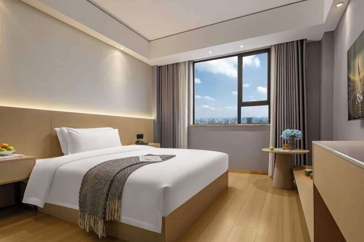 A modern hotel room with a large bed, a small side table, a desk, and a window with a city view.