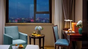 Habitación de hotel con un amplio ventanal que ofrece vistas a la ciudad, un sillón cómodo, una peque ña mesa redonda con un florero y un escritorio con lámpara y adornos.