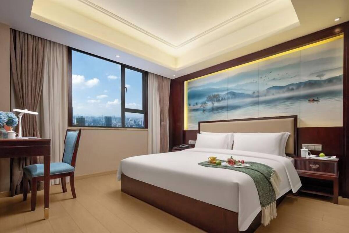 A hotel room with a large bed, a desk with a chair, a nightstand, and a painting on the wall.