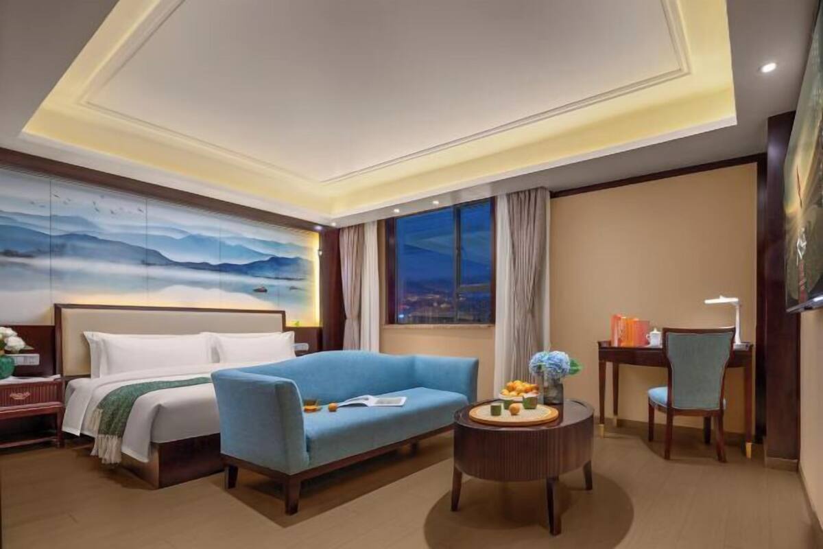 A hotel room with a bed, a blue sofa, a desk with a chair, a round table with a vase, and a painting on the wall.