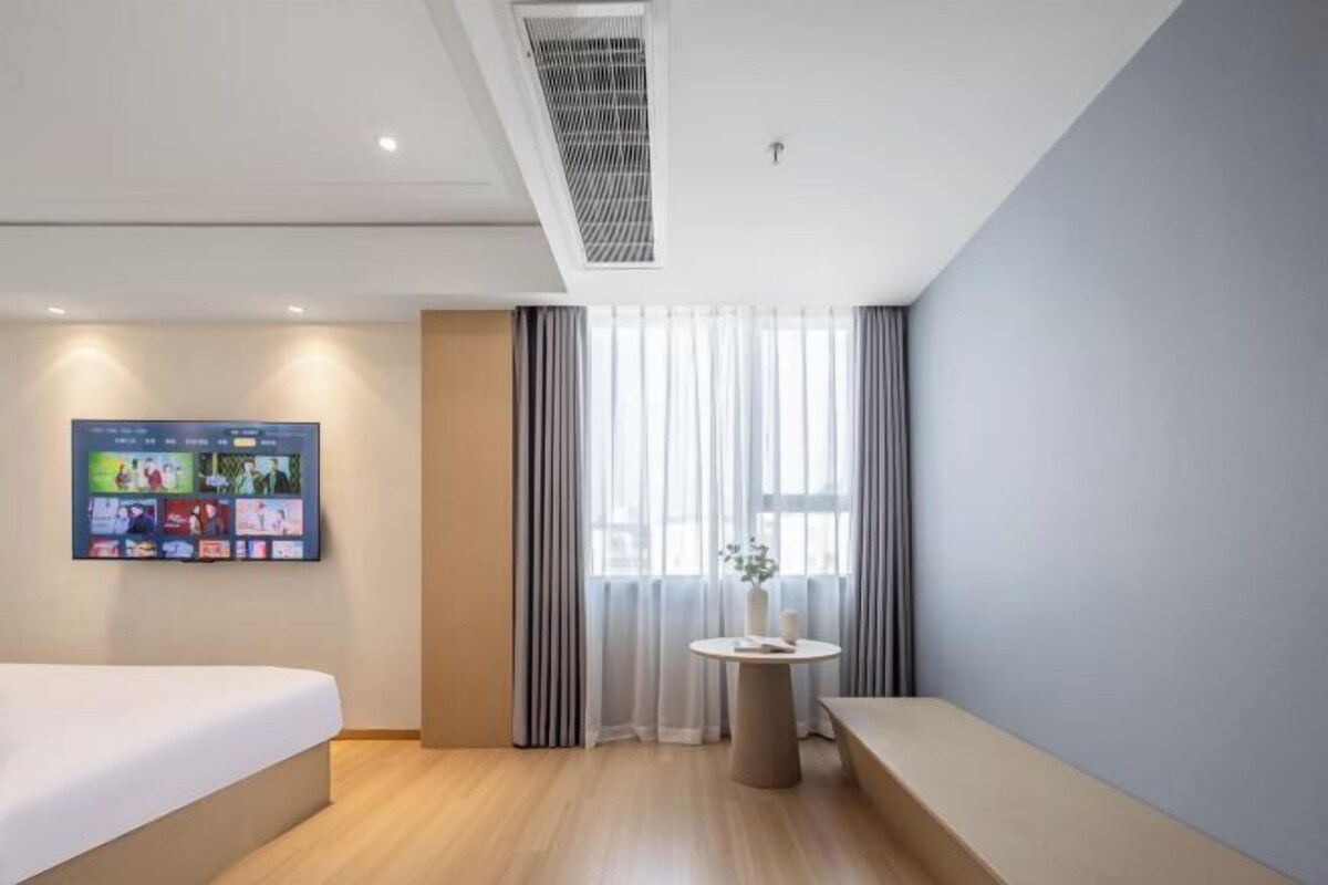 Ein modernes Hotelzimmer mit einem Bett, einem kleinen runden Tisch mit einer Vase und einem großen Fenster mit Vorhängen.