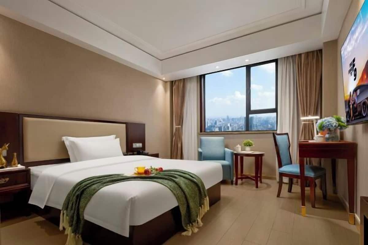 A hotel room with a bed, a desk, a chair, a small table with a plant, and a large window with a city view.