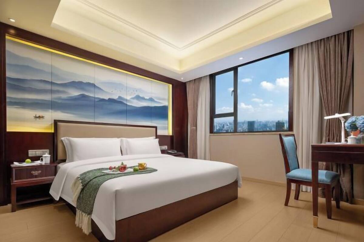 A hotel room with a large bed, a desk with a chair, a nightstand, and a large window offering a city view.