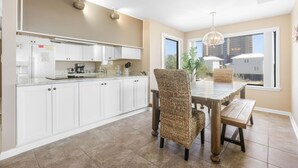 A modern kitchen with white cabinets, a dining area with a wooden table and woven chairs, and a large window with a view of buildings. - Compass Point 201 Pet-friendly for fee Boat Ramp (Gulf Shores)