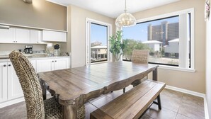 A dining area with a wooden table, bench, and woven chair. There is a window with a view of buildings and a plant on the table. - Compass Point 201 Pet-friendly for fee Boat Ramp (Gulf Shores)