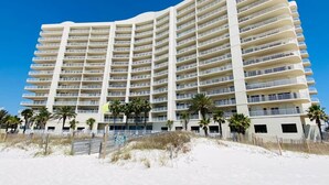 Un hotel de varios pisos con numerosas balconadas, situado en una playa con palmeras y un cielo azul despejado.