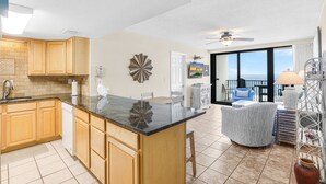 A kitchen with wooden cabinets, a marble island, and a dining area with a view. - Island Winds West 774- Walk to Hangout Beachfront (Gulf Shores)