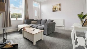 A living room with a grey sofa, a white coffee table, and a window with curtains. - Cosy Green End Retreat (Wakefield)
