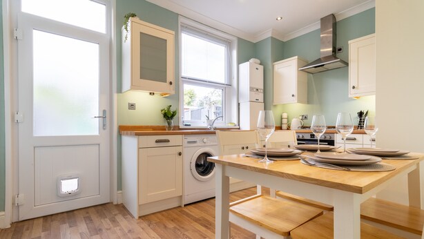 A modern kitchen with white cabinetry, a built-in washing machine, and a dining area with a wooden table and benches. - Cosy Green End Retreat (Wakefield)