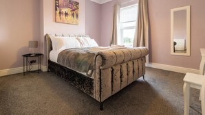 A bedroom with a large bed, a bedside table, a mirror, and a window with curtains. - Cosy Green End Retreat (Wakefield)