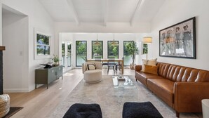 Un salon spacieux avec un canapé en cuir marron, un fauteuil rond blanc et une table basse en verre. On y trouve également un coin repas avec une table et des chaises, ainsi qu’une grande fenêtre donnant sur un paysage verdoyant.