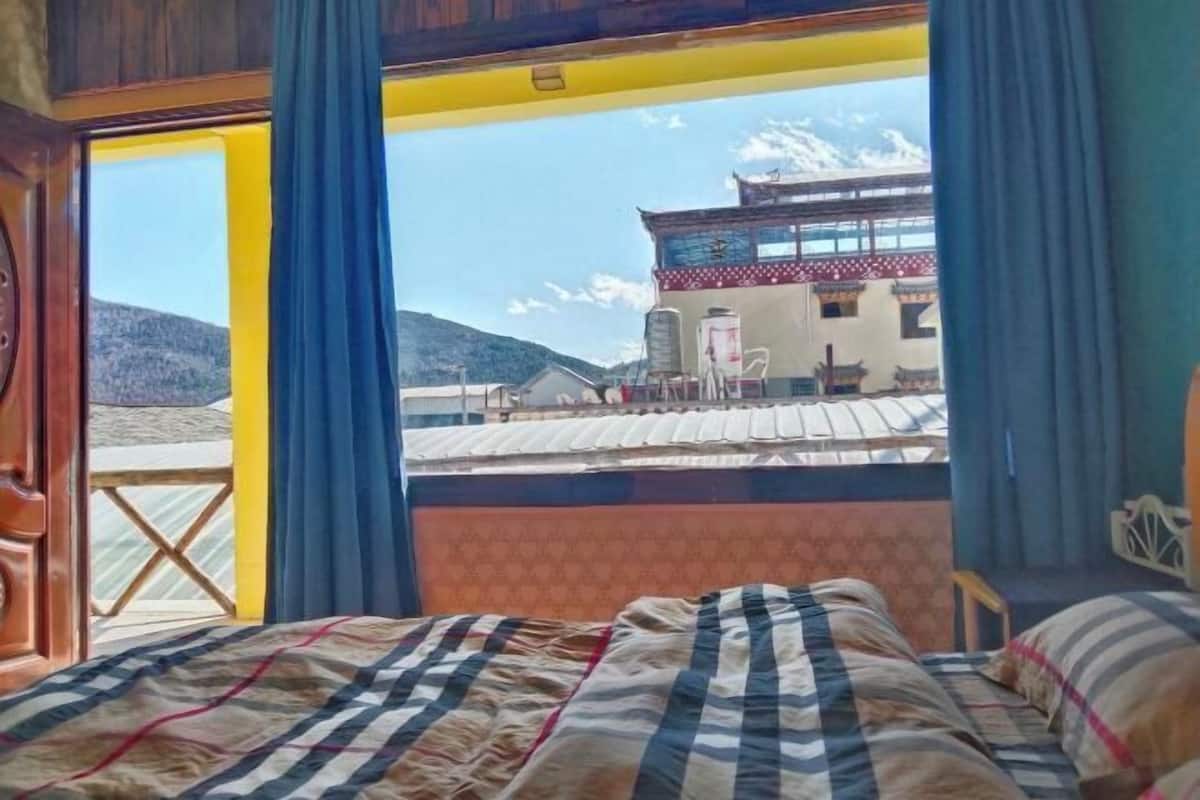 Une chambre avec un lit, une fenêtre avec des rideaux et une vue sur un immeuble et des montagnes.