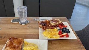 Mesa de desayuno con tostadas, huevos revueltos y fruta, con vista a la playa.