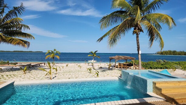 Zona de piscina con vista al océano, rodeada de palmeras y una playa de arena.