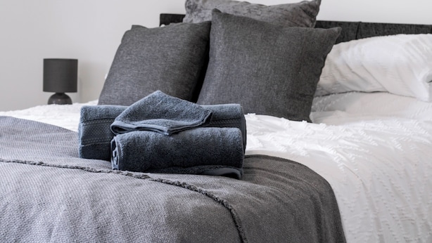 A neatly made bed with grey bedding and pillows, a folded grey towel, and a black bedside lamp. - Lovely Lawns Lane (Carr Gate)