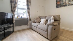 A living room with a flat-screen TV, a sofa with plaid cushions, a window with plaid curtains, and a cityscape painting on the wall. - Lovely Lawns Lane (Carr Gate)