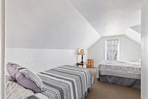 A small, white, attic-style bedroom with two beds, a window with blinds, a lamp, and a small table.