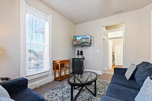 A living room with a sofa, a glass coffee table, a TV mounted on the wall, and a window with curtains.