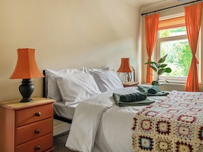Ein ordentlich bezogenes Bett mit weißen Leinen, zwei Nachttischlampen, einer Zimmerpflanze und einer gehäkelten Tagesdecke.