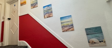Una escalera con peldaños rojos que conduce a una puerta blanca, decorada con cuatro obras de arte con motivos playeros.