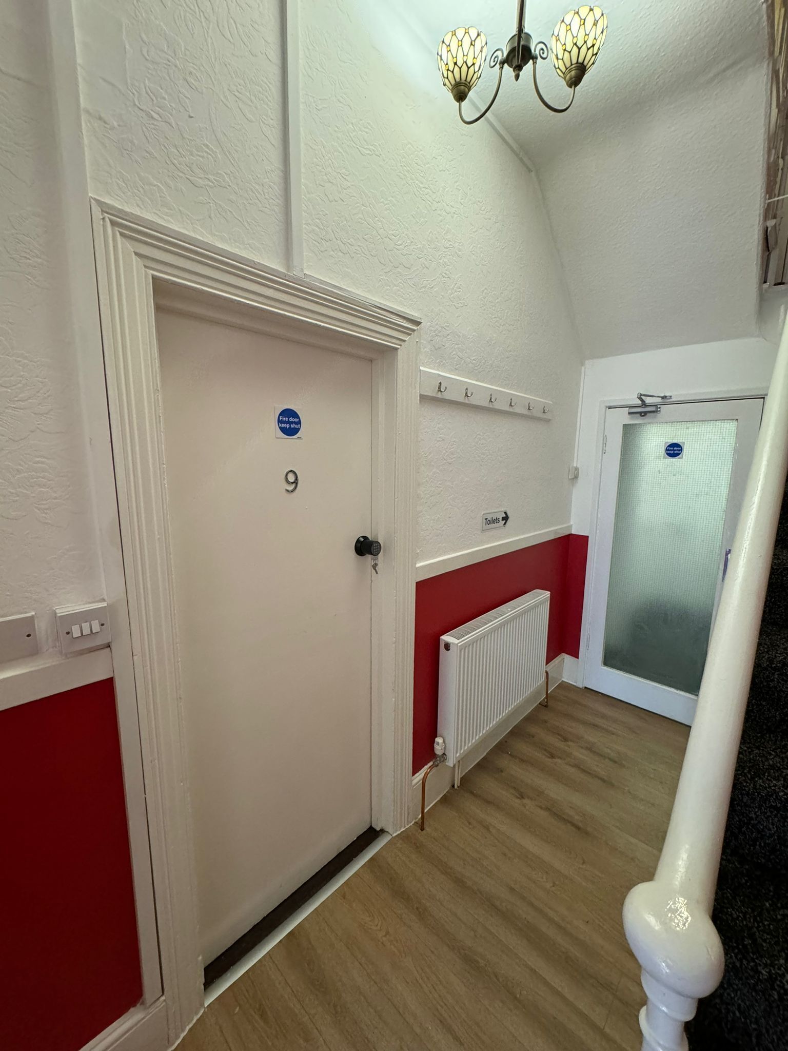 Un couloir avec une porte blanche portant le numéro 9, un radiateur et une porte vitrée avec une pancarte.