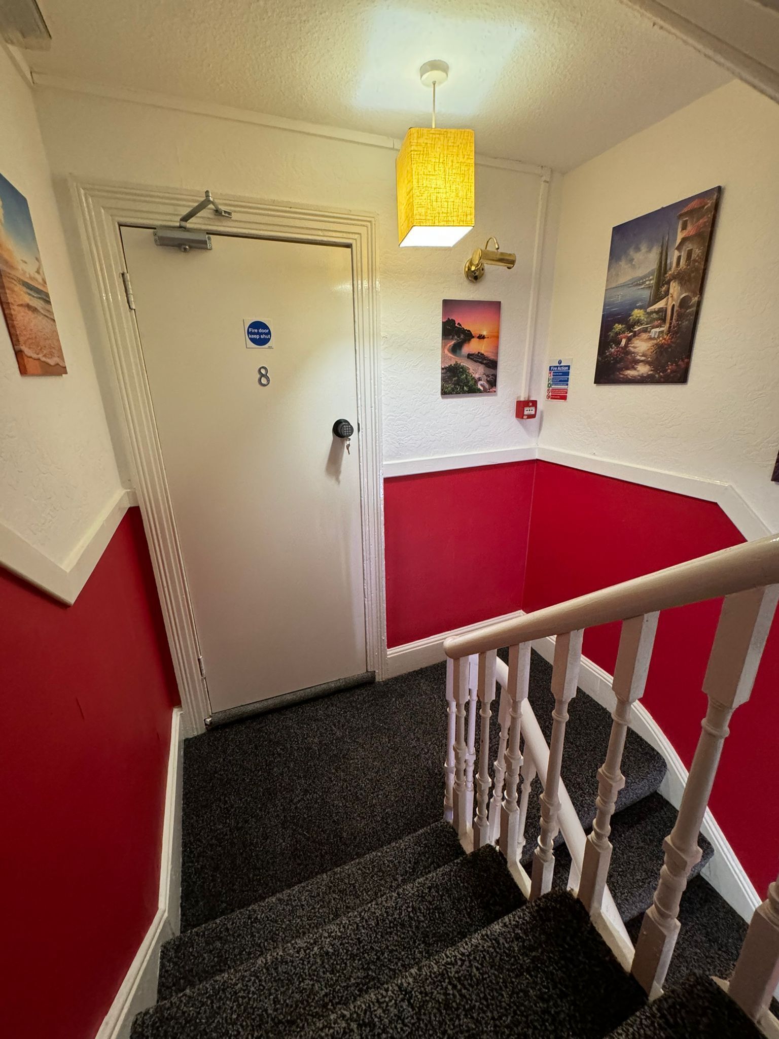 Eine Treppe mit roter und weißer Wand, eine Tür mit Brandschutznachweis, eine Hängelampe und ein Gemälde an der Wand.