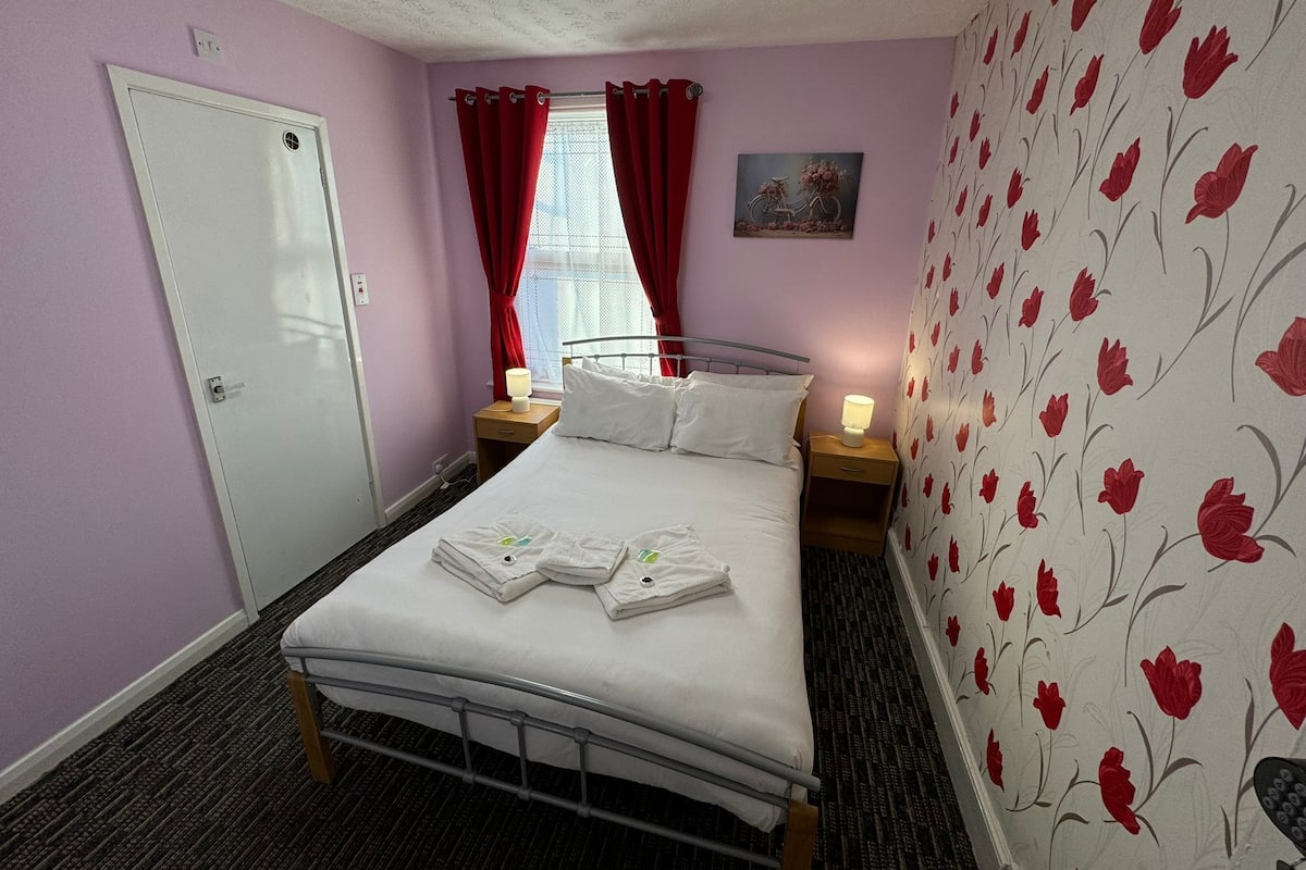 Une chambre à coucher avec un lit, deux tables de chevet, une fenêtre avec des rideaux rouges, une porte et un papier peint à motifs floraux sur un des murs.