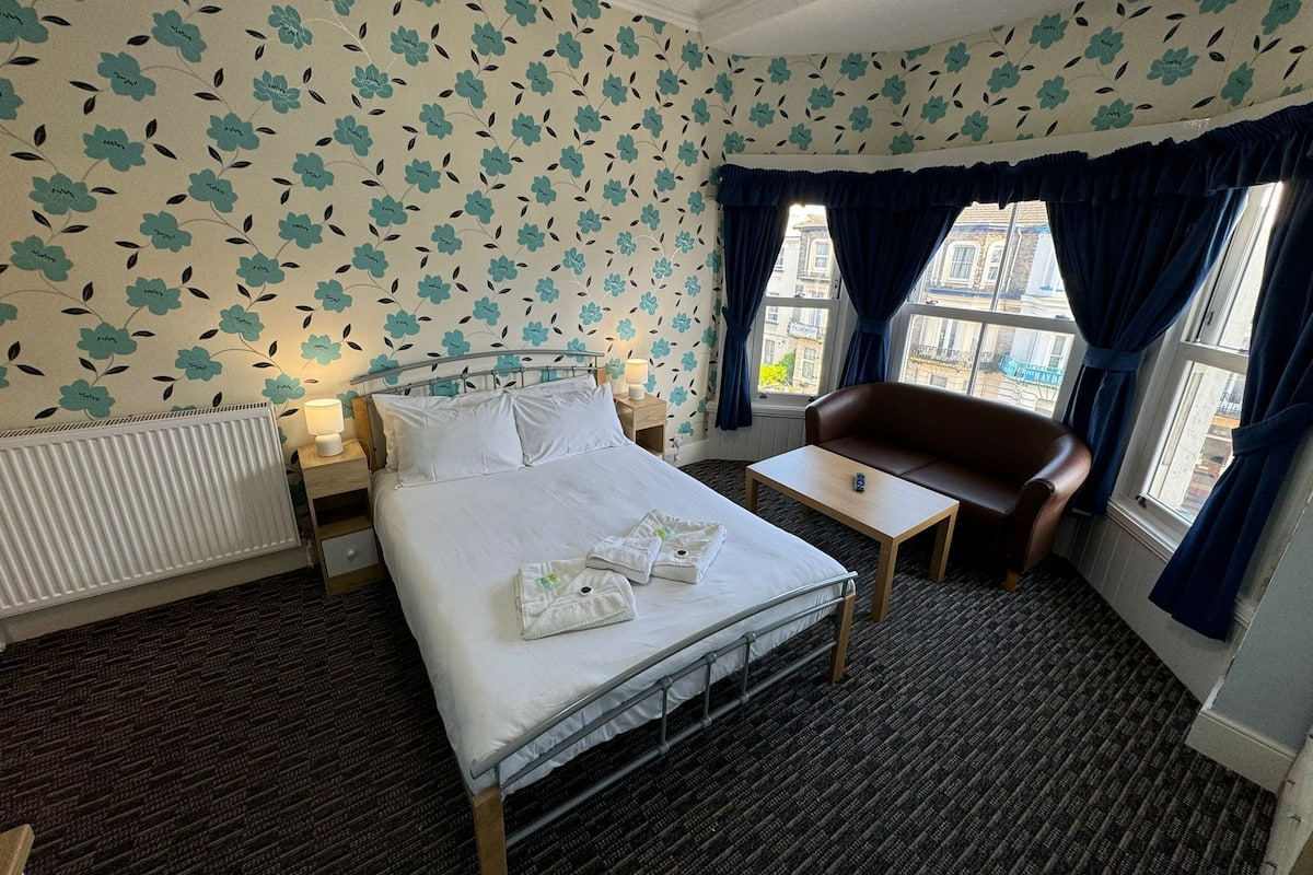 Une chambre d’hôtel avec un lit, une petite table, un canapé et une fenêtre avec des rideaux.