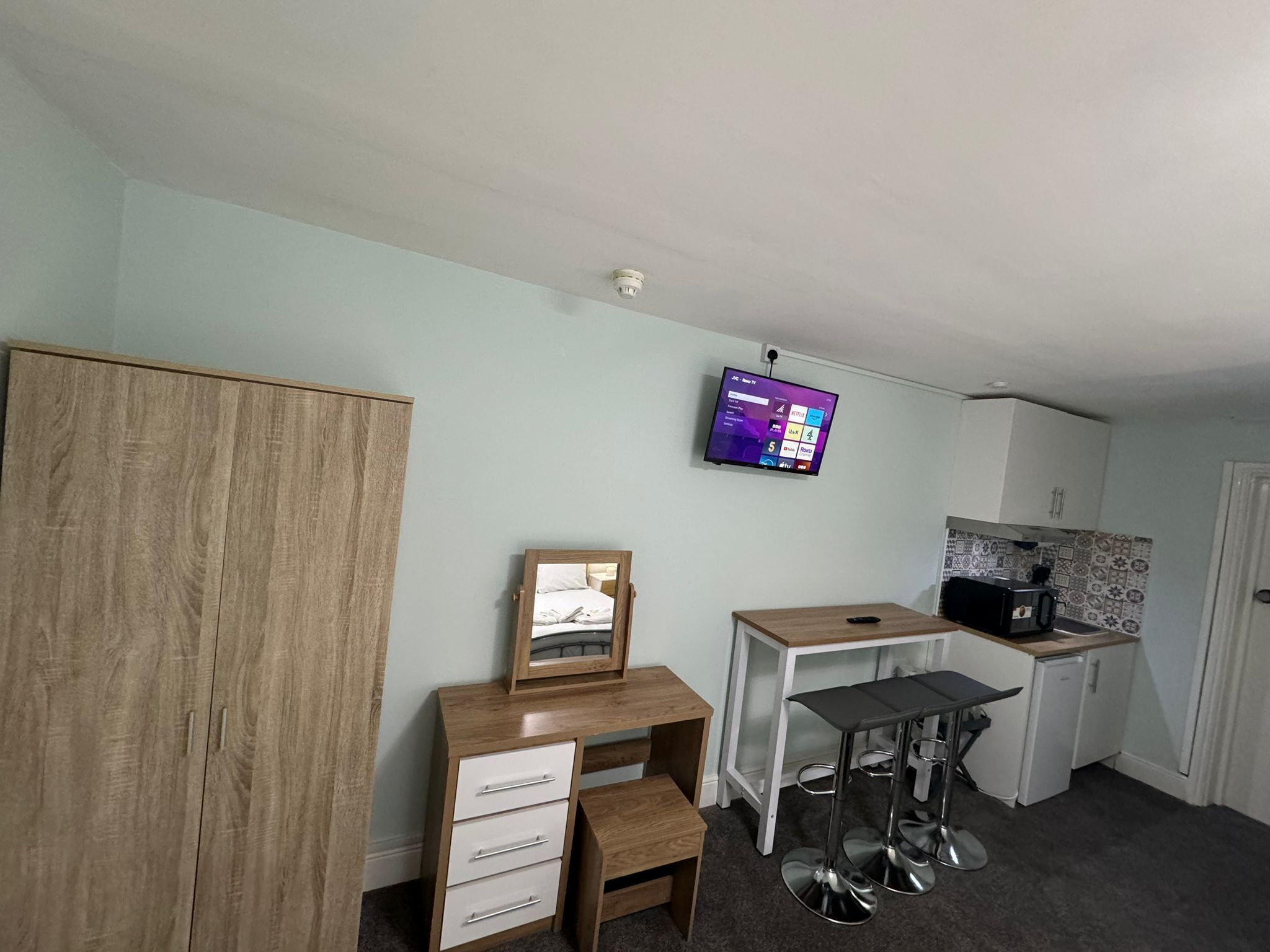 A compact room with a wooden wardrobe, a desk with drawers, a small stool, a mounted TV, and a kitchenette area.