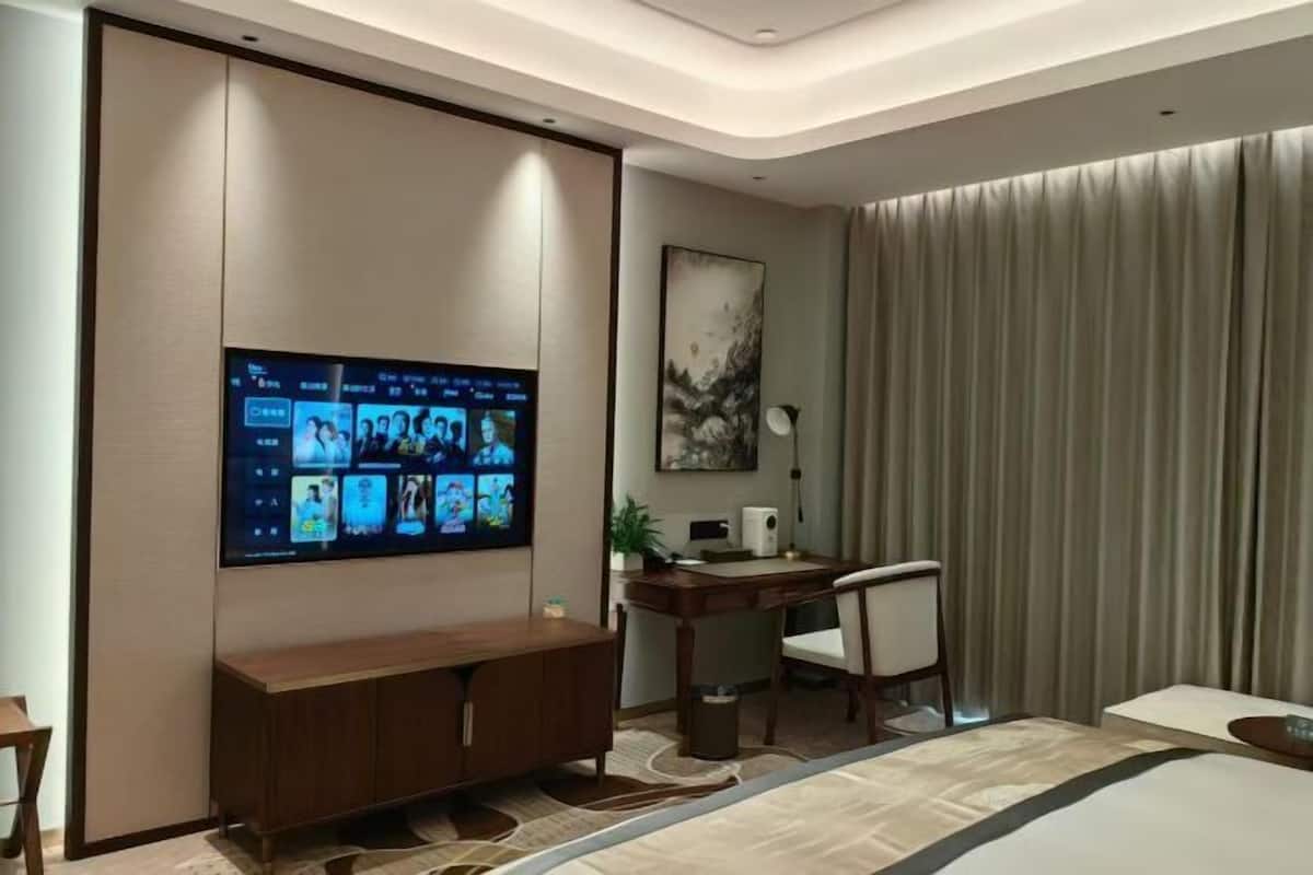 Habitación de hotel moderna con una gran televisión de pantalla plana montada en la pared, un escritorio de madera con una silla, una cama con colcha a rayas color beige y gris, y un amplio ventanal con cortinas.
