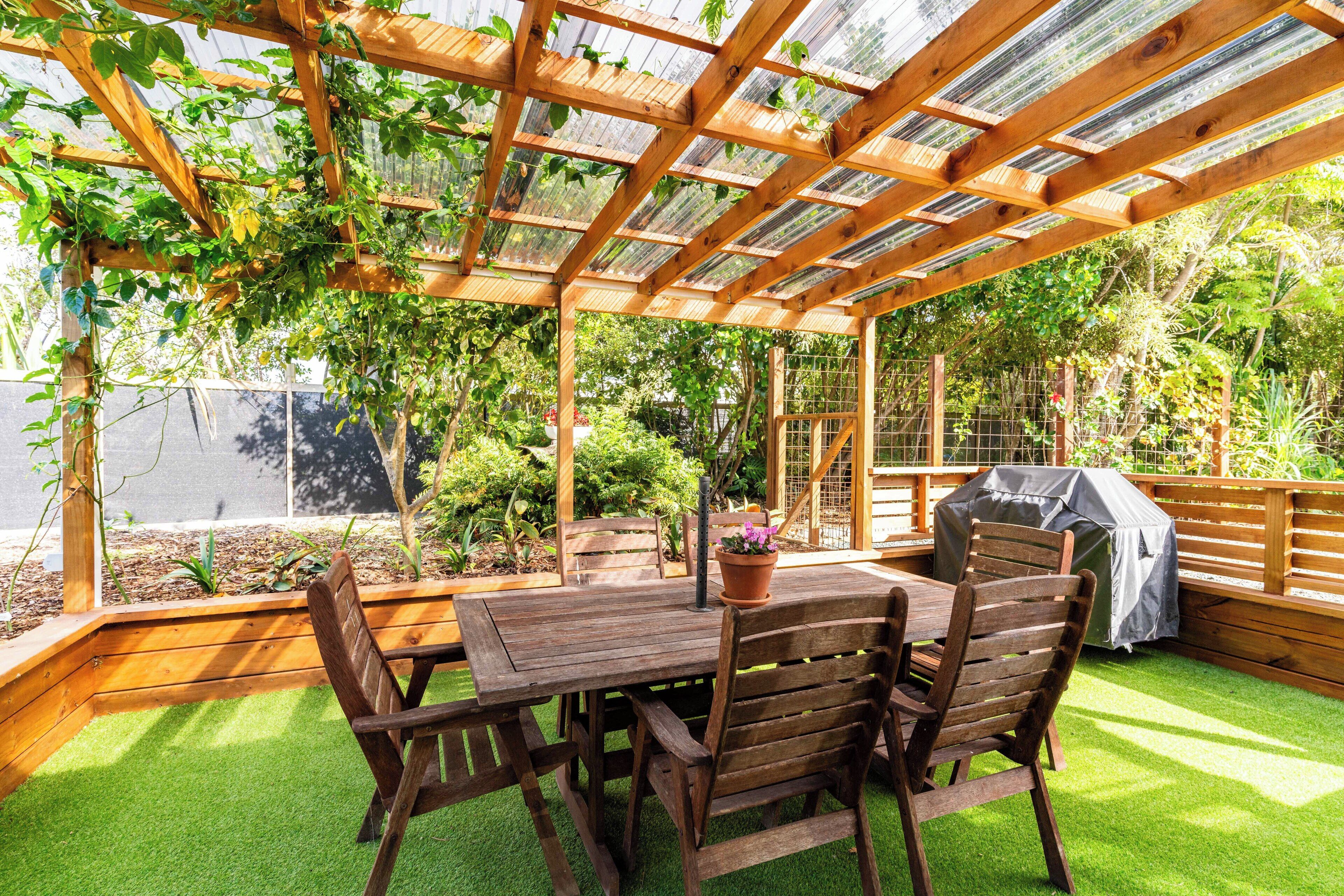 Eine hölzerne Pergola mit Tisch und Stühlen, umgeben von grünem Gras und Pflanzen.