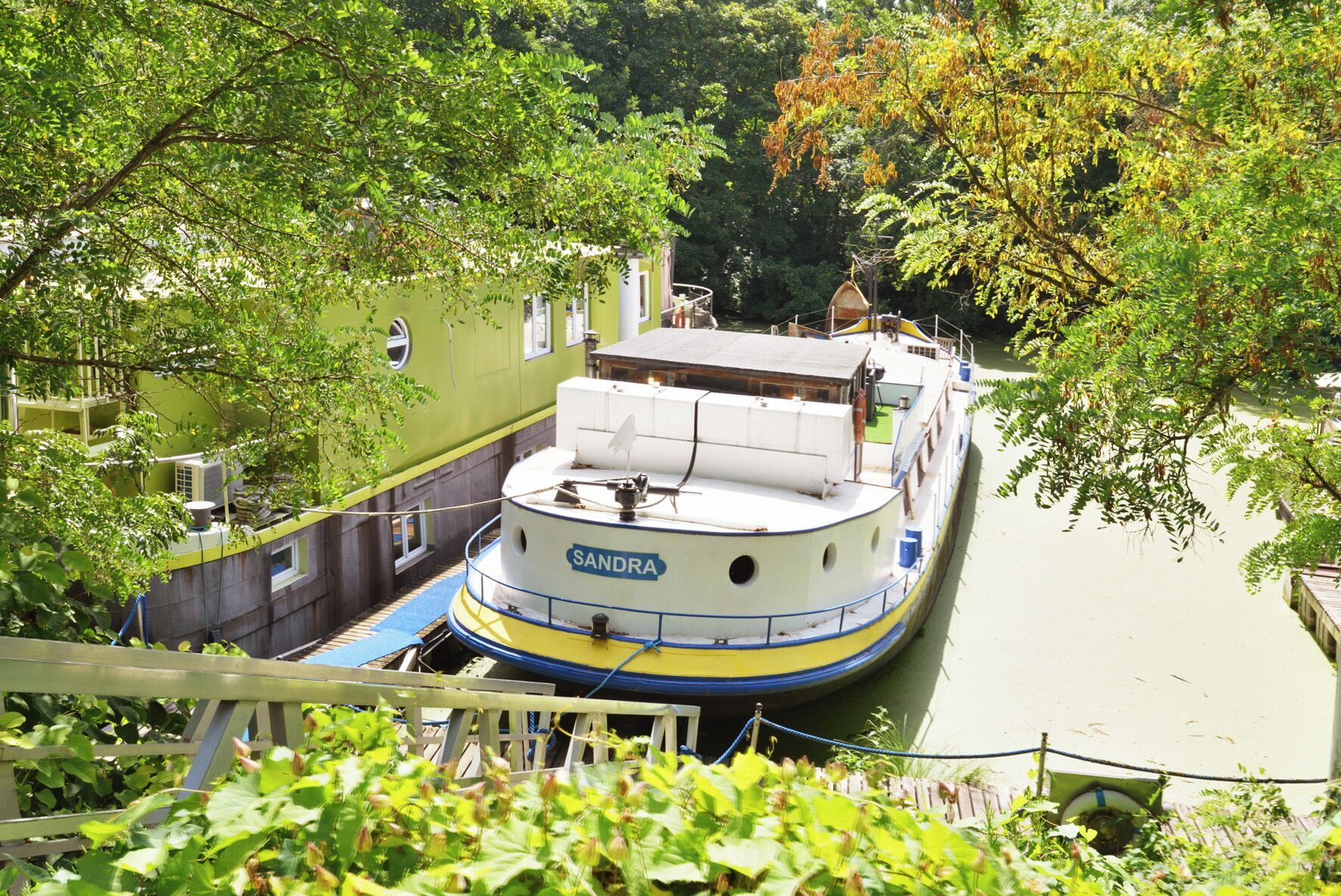 Ein schmaler Kanal mit einem Boot namens 'SANDRA', das an einem grünen Gebäude mit Fenstern anlegt.