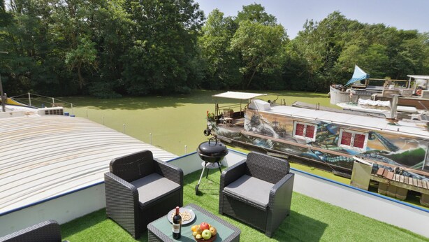 A rooftop terrace with wicker furniture, a barbecue, and a view of a river with a boat. - Peniche Refuge Seine (Vigneux-sur-Seine)