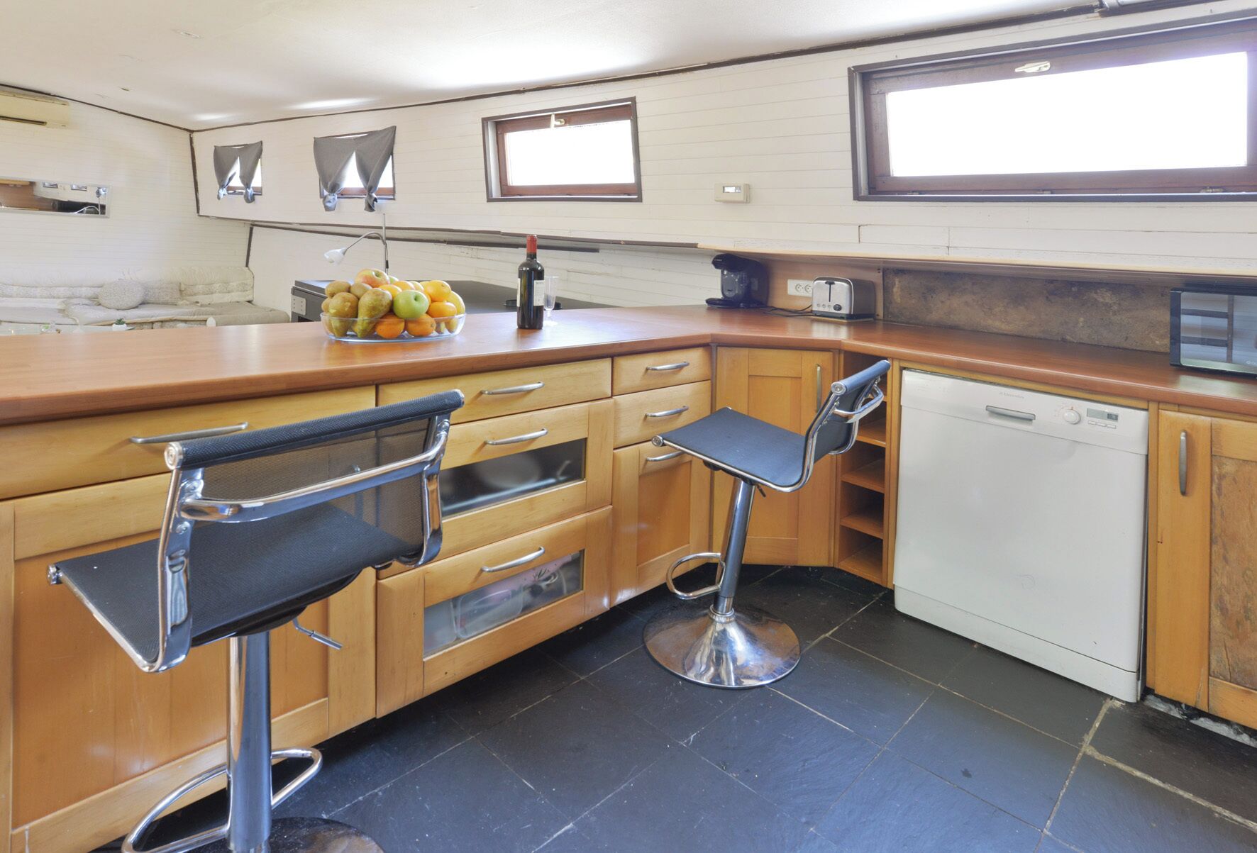 Une cuisine compacte avec des meubles en bois, un sol carrelé noir, un lave-vaisselle blanc et deux tabourets de bar.
