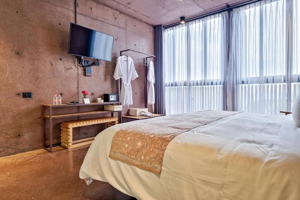 A hotel room with a large bed, a TV mounted on the wall, a bedside table with a lamp, and a window with sheer curtains.