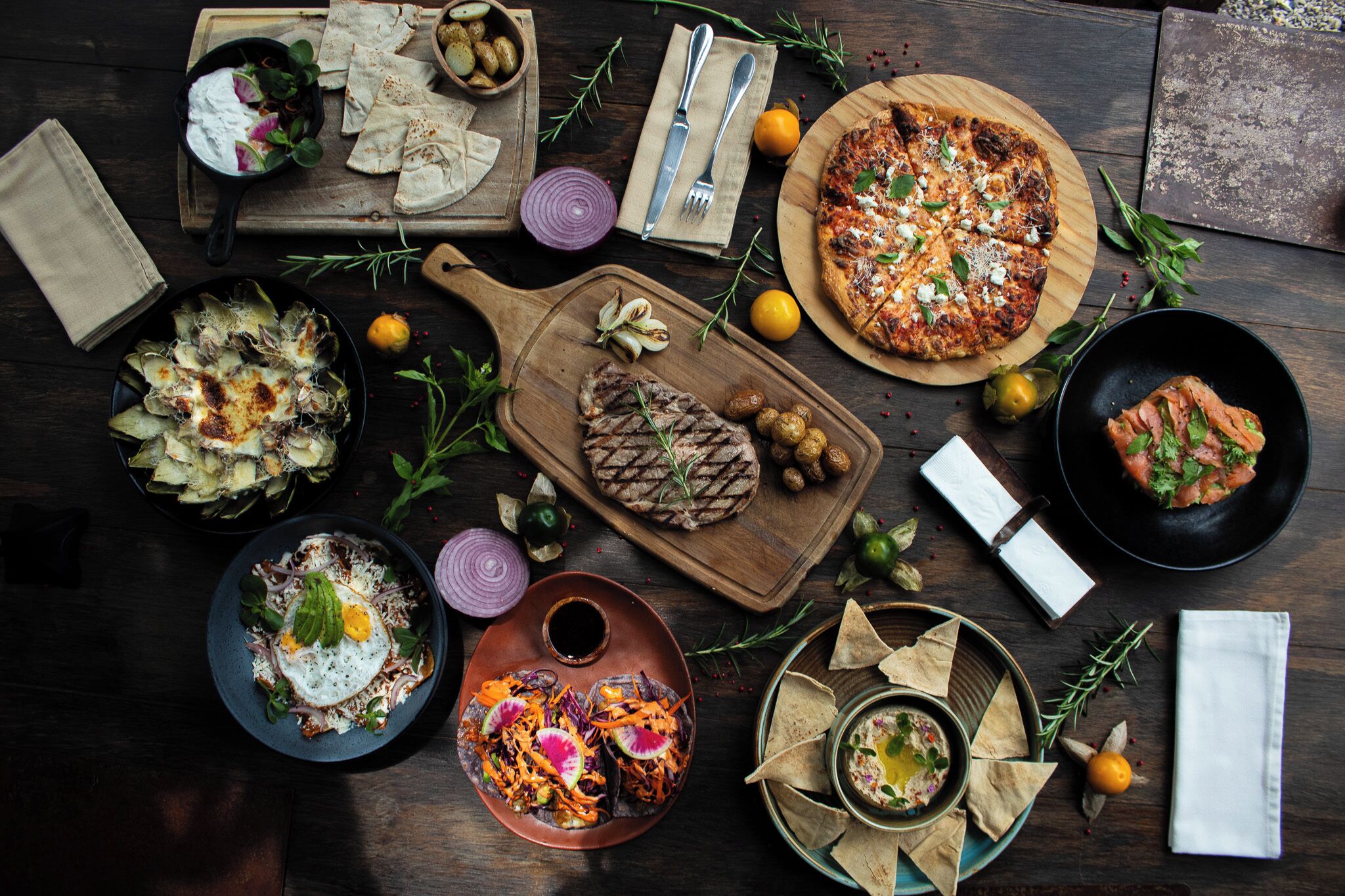 A variety of dishes including a grilled steak, a pizza, and a salad, arranged on a wooden table.