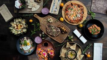 Forskellige retter, heriblandt en grillet steak, pizza og en salat, anrettet på et træbord.