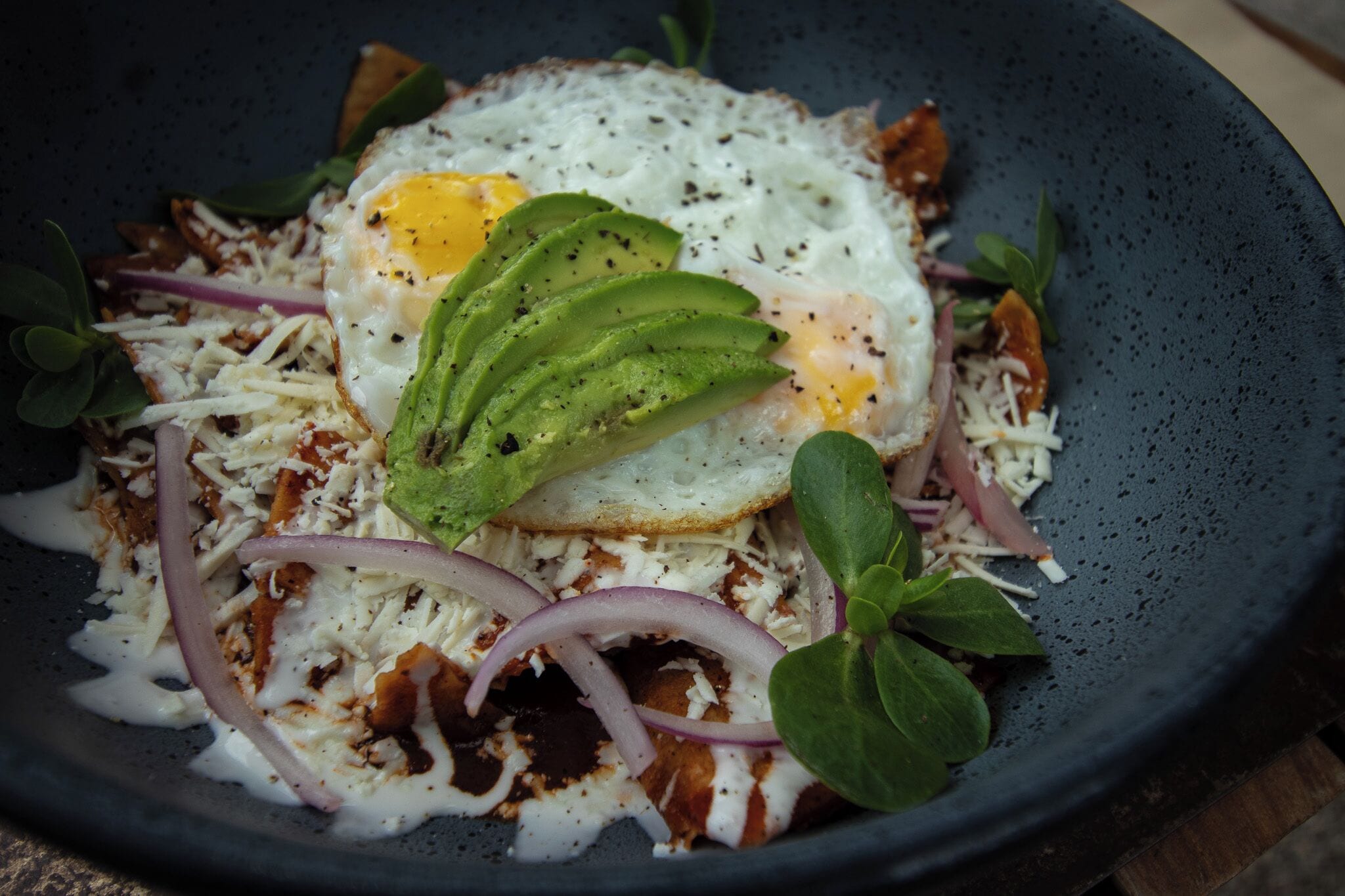 Un plat composé d’un œuf au plat, de tranches d’avocat, de fromage râpé et d’oignon rouge, le tout servi dans une assiette foncée.