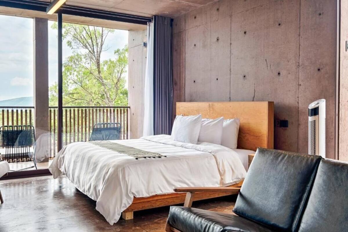 A bedroom with a large bed, a wooden headboard, a black leather sofa, and a view of trees through large windows.
