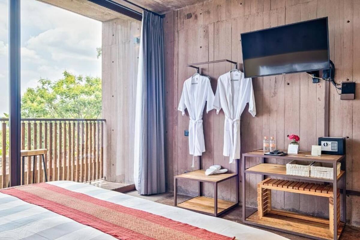 A room with a bed, a TV mounted on the wall, a small table with a vase and bottles, and a balcony with a wooden railing.