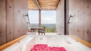 A spa room with a large bathtub filled with rose petals, wooden flooring, and a balcony with a table and chairs.