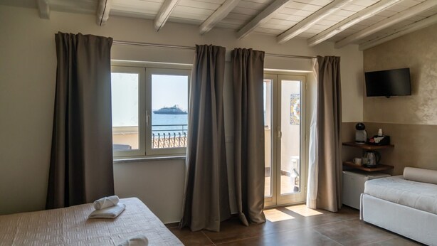 A hotel room with a bed, a sofa, a TV, and a balcony with curtains. - giardini inn arte (Giardini Naxos)
