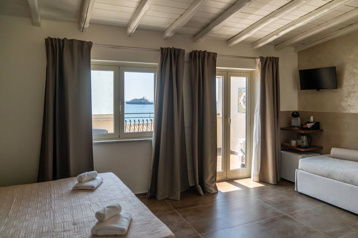 Ein Hotelzimmer mit Bett, einer Couch, einem Fernseher und einem Balkon mit Vorhängen.