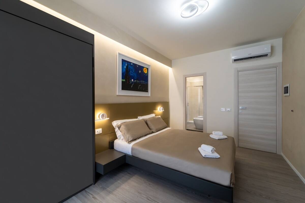 Ein modernes Hotelzimmer mit Bett, Nachttischlampen, einem Himmel-Gemälde, einem Badezimmerspiegel und einer Klimaanlage.
