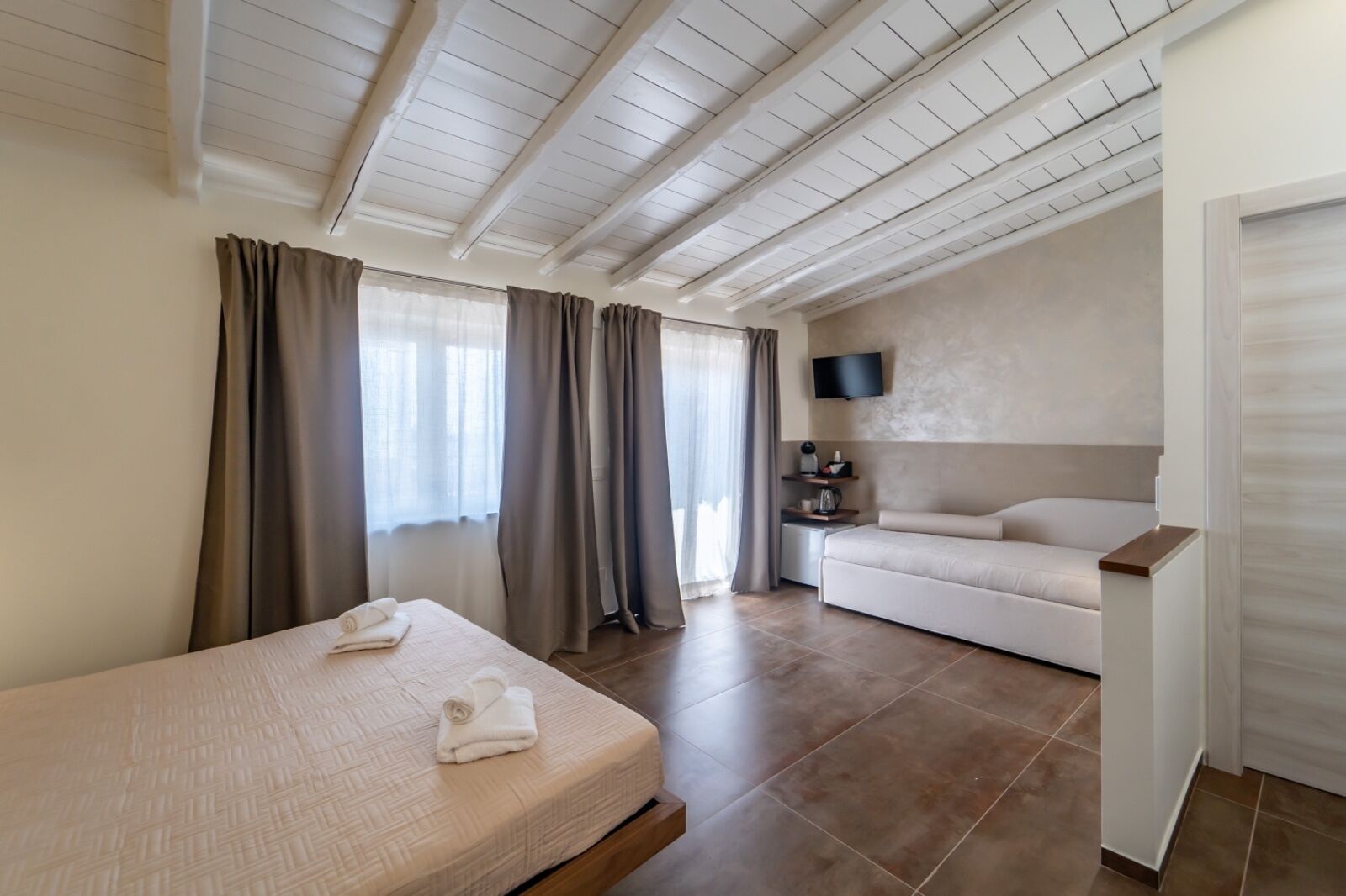 A hotel room with a bed, a sofa, a TV mounted on the wall, and a small table with a kettle.