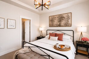 A bedroom with a bed, bedside tables, a nightstand, a floor lamp, a ceiling light fixture, a door, and a framed picture on the wall.