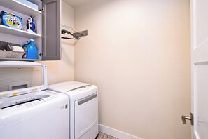 Zona de lavandería con lavadora y secadora, estantes con productos de limpieza y una puerta a la derecha.